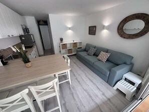 A modern kitchen with white cabinetry, a dining table with chairs, a grey sofa with patterned cushions, and a small white stool. - 2-room apartment with pool, tennis and private parking 50m from the beach (Port Camargue)