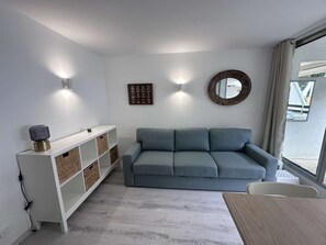 A modern living room with a grey sofa, a white shelving unit, and a round mirror on the wall. - 2-room apartment with pool, tennis and private parking 50m from the beach (Port Camargue)