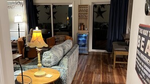 Ein gemütliches Wohnzimmer mit einem gemusterten Sofa, einem kleinen Holztisch und einer Lampe. Ein Bücherregal mit Dekorationselementen und ein Schild an der Wand sind ebenfalls zu sehen.