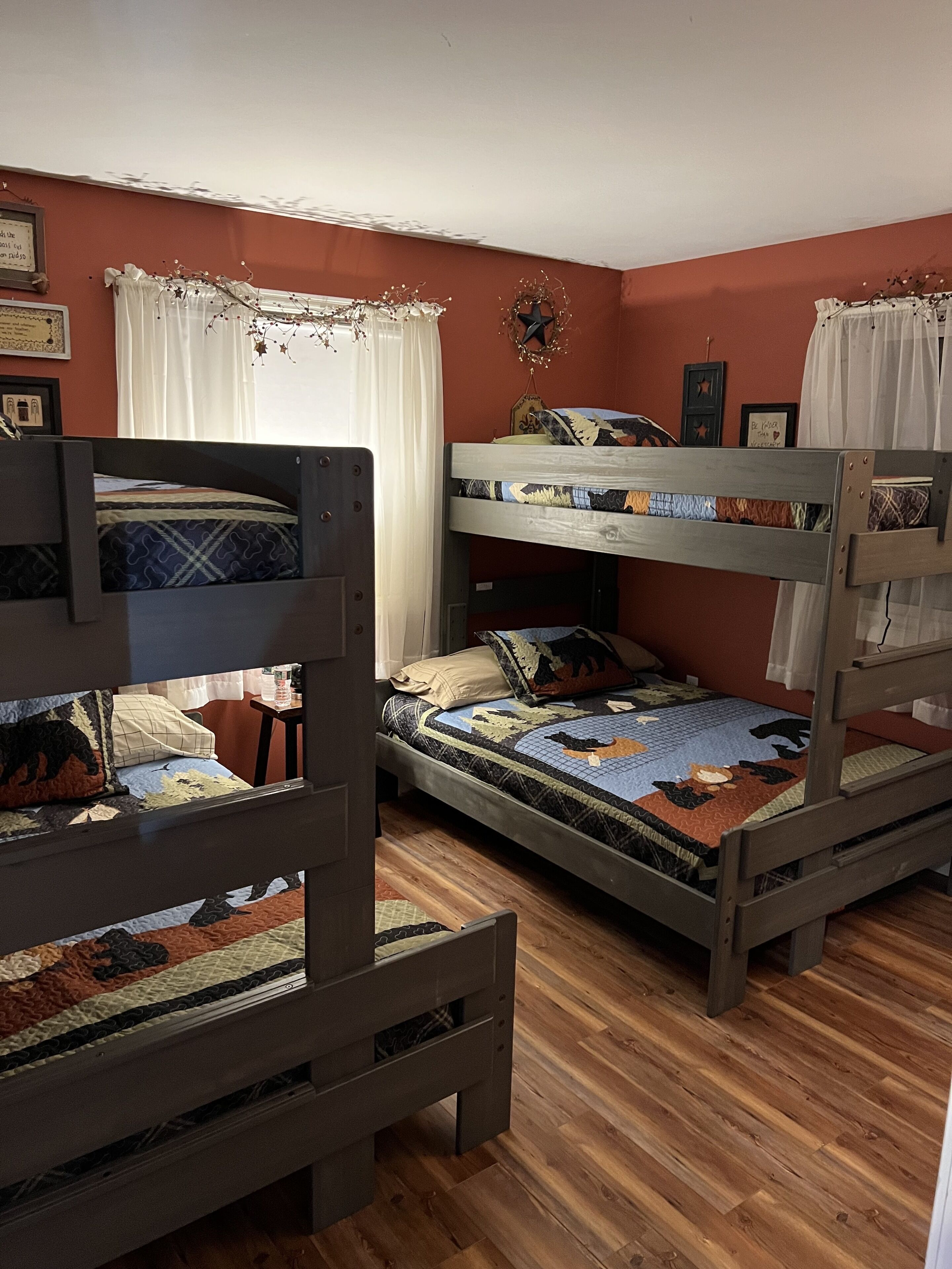 A room with three bunk beds, wooden flooring, and a window with white curtains.