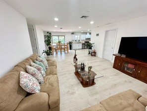 A living room with a sofa, coffee table, TV stand, and a kitchen in the background.