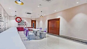 A spacious room with a long white table, round tables, and chairs, a large wooden wardrobe, and a wall with a motivational quote. - Hotel Veridian (Lucknow)