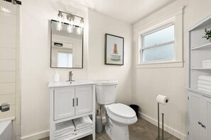 A modern bathroom with a white sink, toilet, and vanity. There is a window providing natural light and a framed picture on the wall. - Walk to Todos Santos | Historic Downtown Delight (Concord)