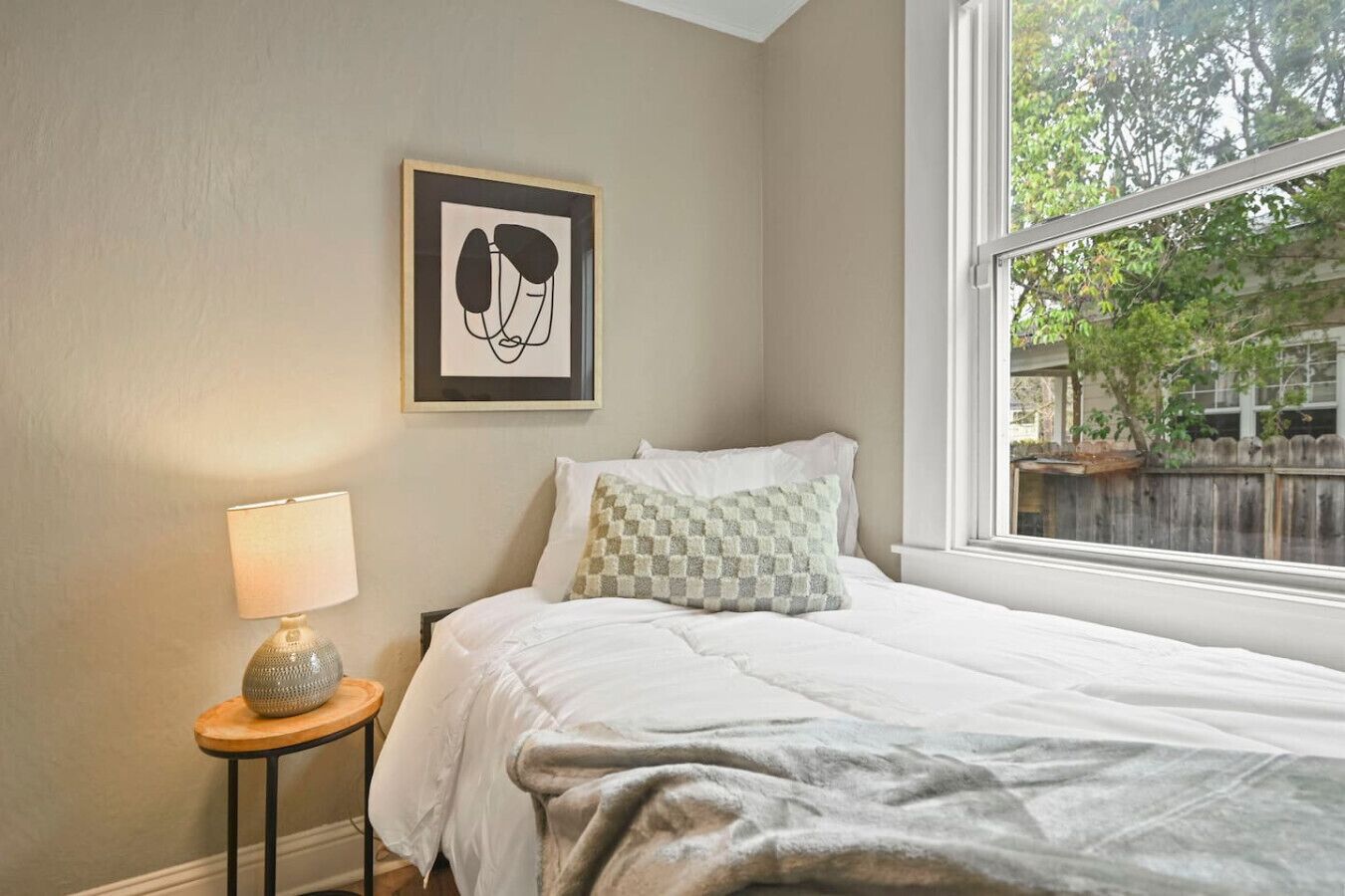 Un dormitorio con cama, lámpara de noche, una obra de arte enmarcada y ventana con vista a árboles.