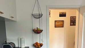 Eine Küche mit schwarzem Arbeitsplatz, einem hängenden Obstkorb und einer Tür zu einem Korridor mit einem gemusterten Teppich.
