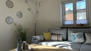 Ein modernes Wohnzimmer mit grauem Sofa, Wandkunst, einer stehenden Stehlampe und einem Fenster mit Blick auf Gebäude.