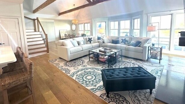 Un vaste salon comprenant un grand canapé d’angle, une table basse et un pouf foncé. Un escalier avec des rampes en bois mène à l’étage supérieur. La pièce est dotée de grandes fenêtres qui laissent entrer la lumière naturelle.