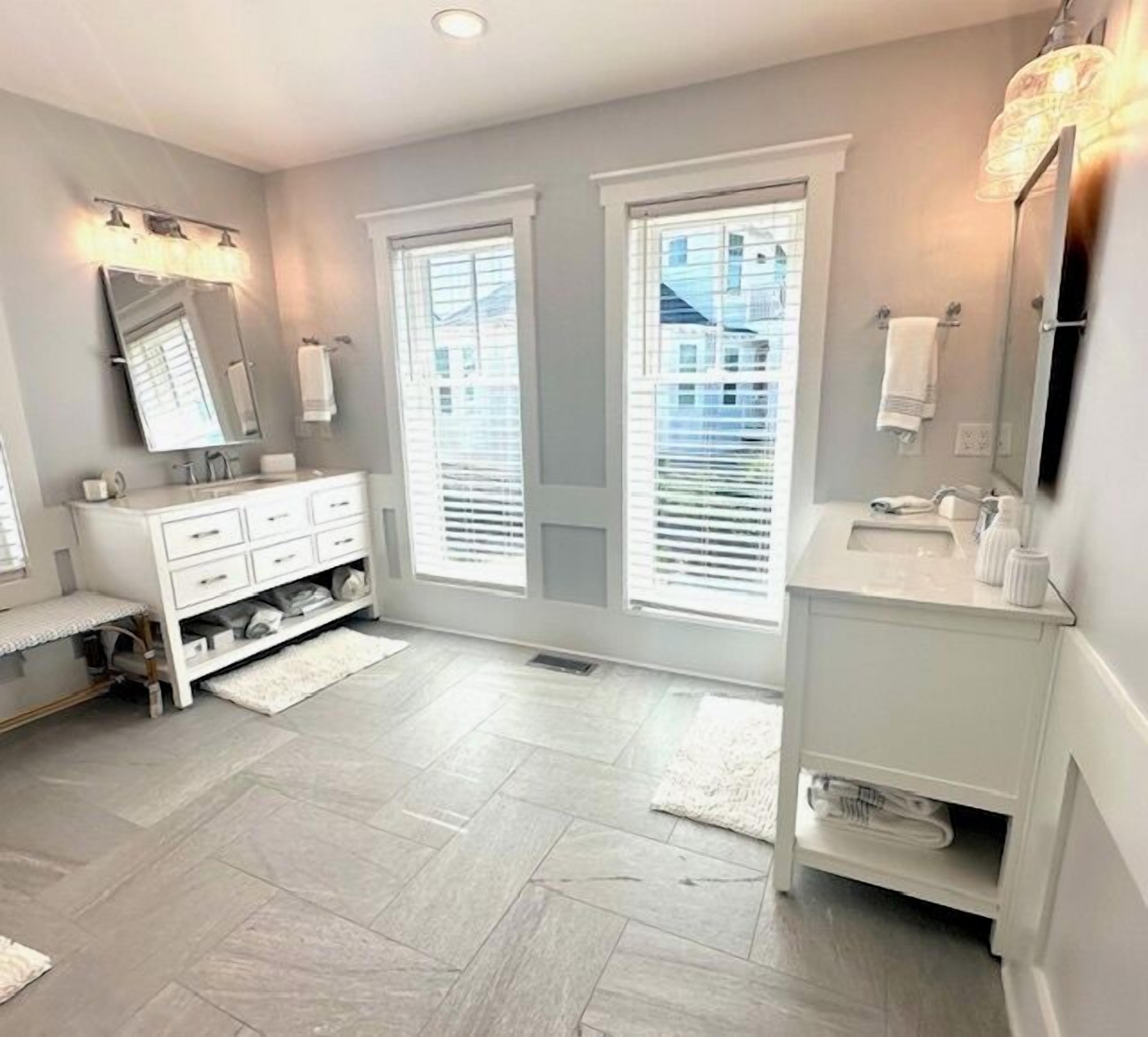 Une salle de bain moderne avec un meuble-lavabo blanc, un grand miroir et deux fenêtres équipées de stores.