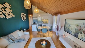 Una sala moderna con un sofá, una mesita de centro y un comedor con sillas. Se observa una cocina al fondo con armarios blancos y una mesa de comedor.