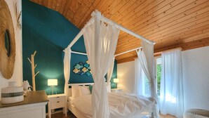 Un dormitorio con cama con dosel, una mesita de noche con lámpara, un adorno en la pared y vistas al verdor exterior.