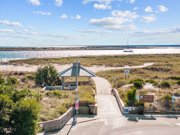 Un acceso a la playa con zona de espera techada, un letrero que indica 'no se admiten perros' y un cartel de acceso público regional a la playa.