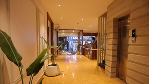 A hotel corridor with a polished floor, potted plants, an elevator, and a visible store in the background.