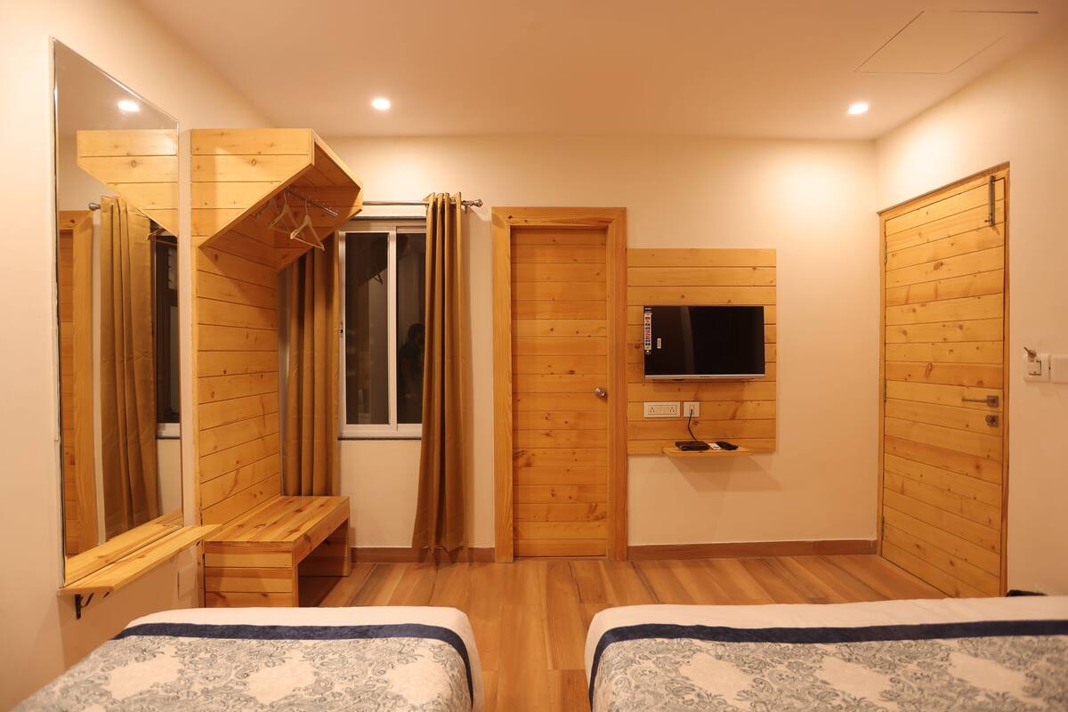 Un dormitorio con dos camas, un banco de madera, un televisor montado en la pared y una ventana con cortinas.