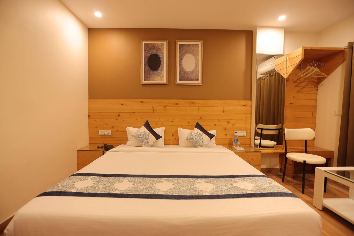 Un dormitorio ordenado con cabecera de madera, cama con colcha estampada, dos obras de arte colgadas en la pared, un escritorio con una silla y una zona de ducha con toallero.