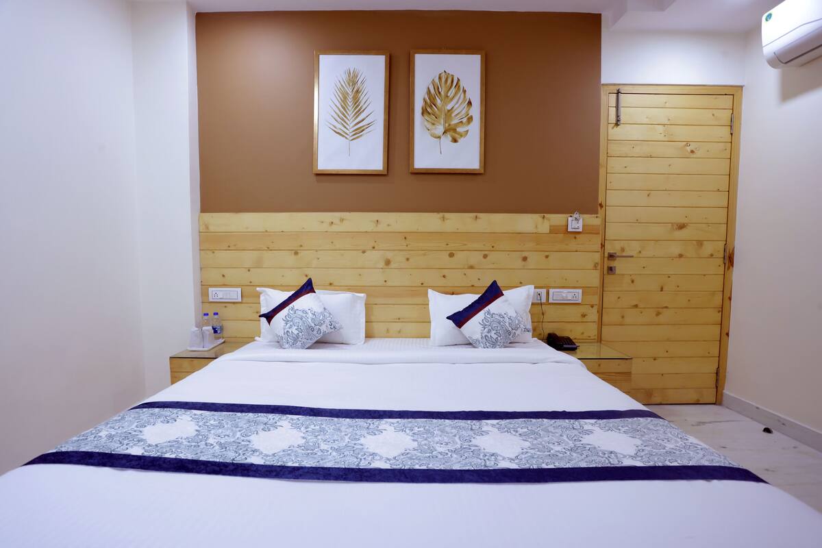 Habitación de hotel con cabecera de madera, cama con ropa de cama blanca, dos obras de arte con hojas enmarcadas en la pared y una puerta de madera.