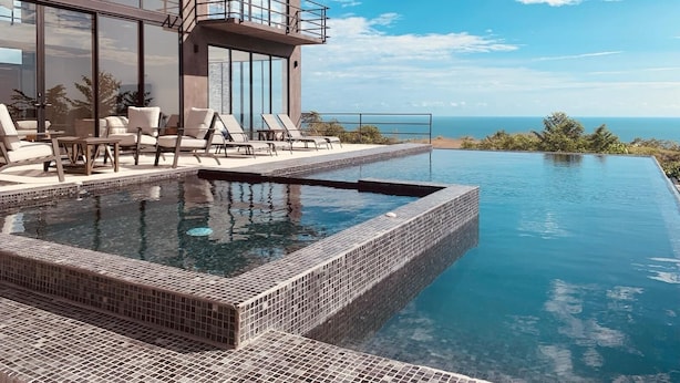 Une piscine à débordement moderne offrant une vue dégagée sur l’océan, une terrasse en bois avec des chaises longues et un grand bâtiment en verre doté d’un balcon.