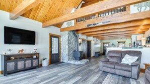 A spacious living area with a stone fireplace, a large flat-screen TV, and a high ceiling with exposed beams. - Lakefront New Hampshire Hideaway w/ Deck & Views (Errol)