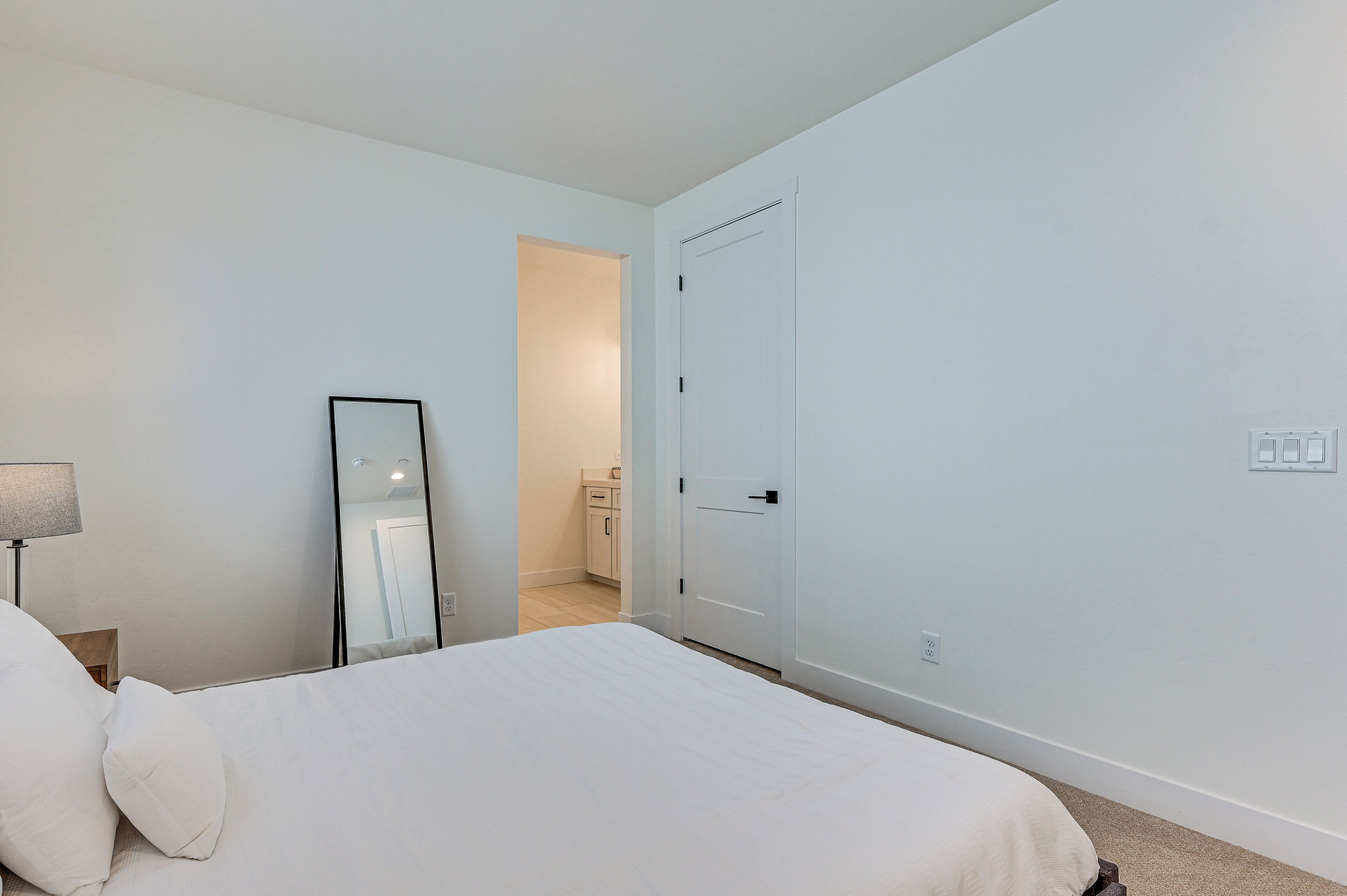 A bedroom with a bed, a bedside lamp, a mirror, and a door leading to a bathroom.