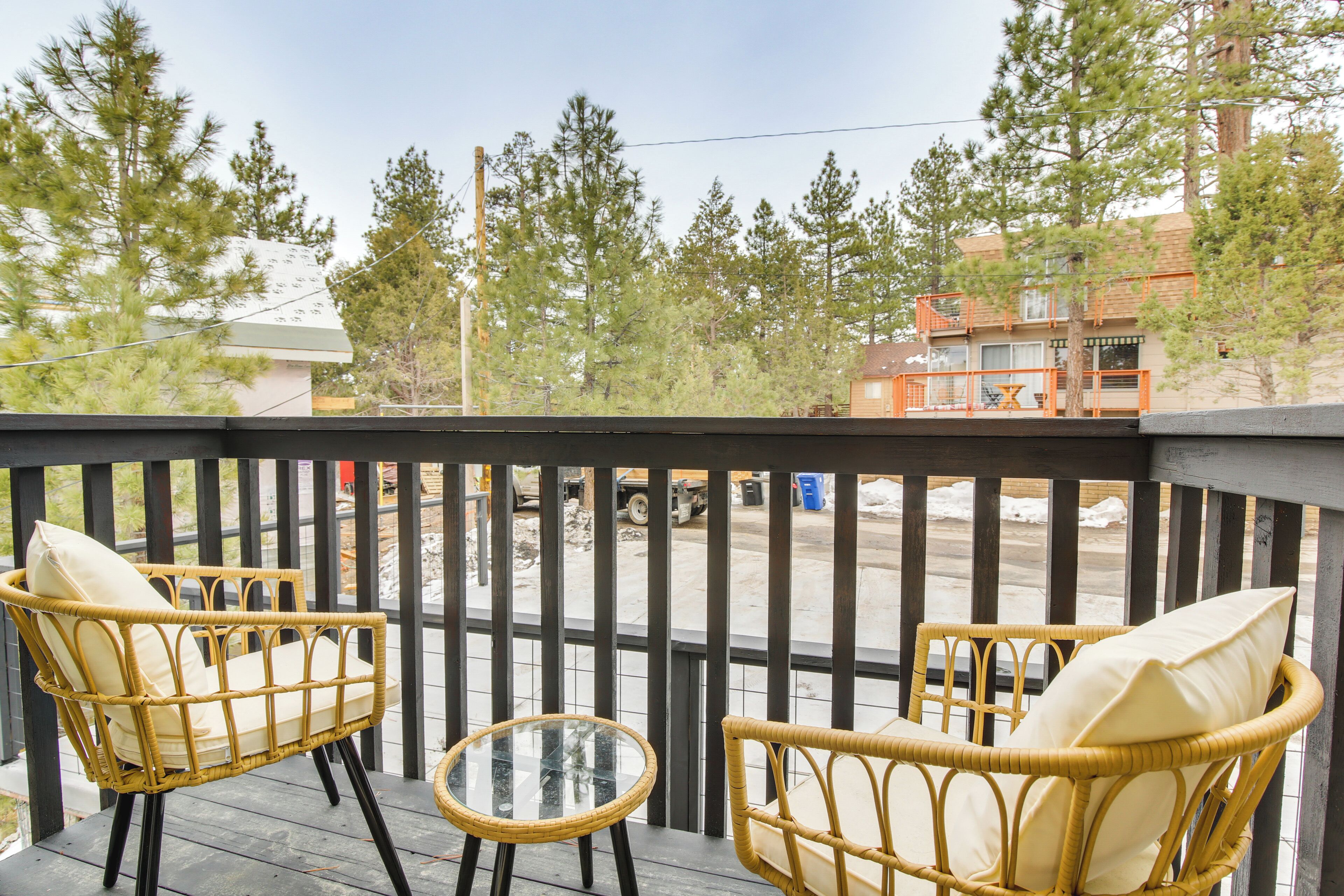 Una terraza de madera con sillas de mimbre y una mesa de vidrio, con vista a una calle con edificios y árboles.