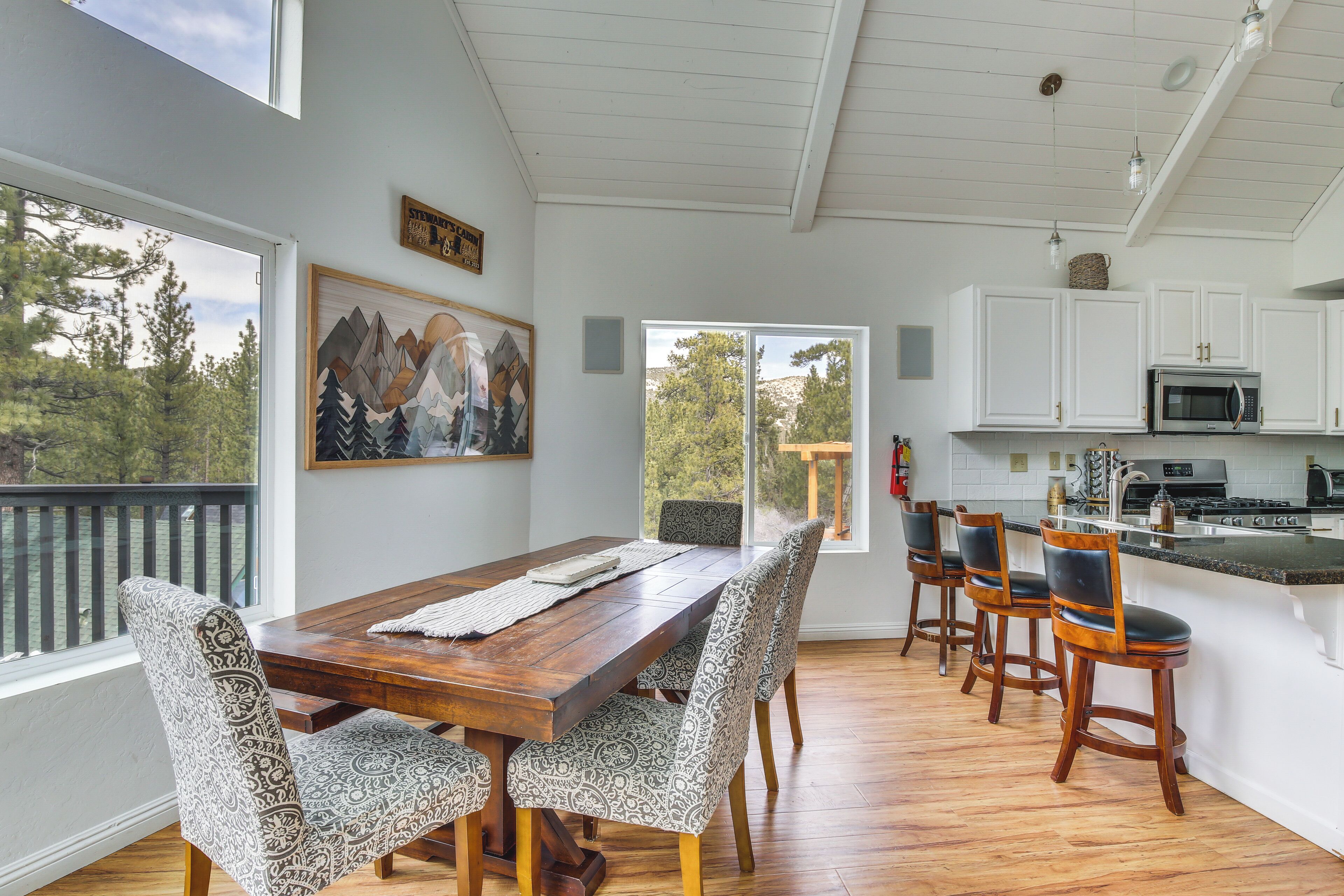 A spacious dining area with a wooden table and patterned chairs, a kitchen with white cabinets, and a view of trees through large windows.