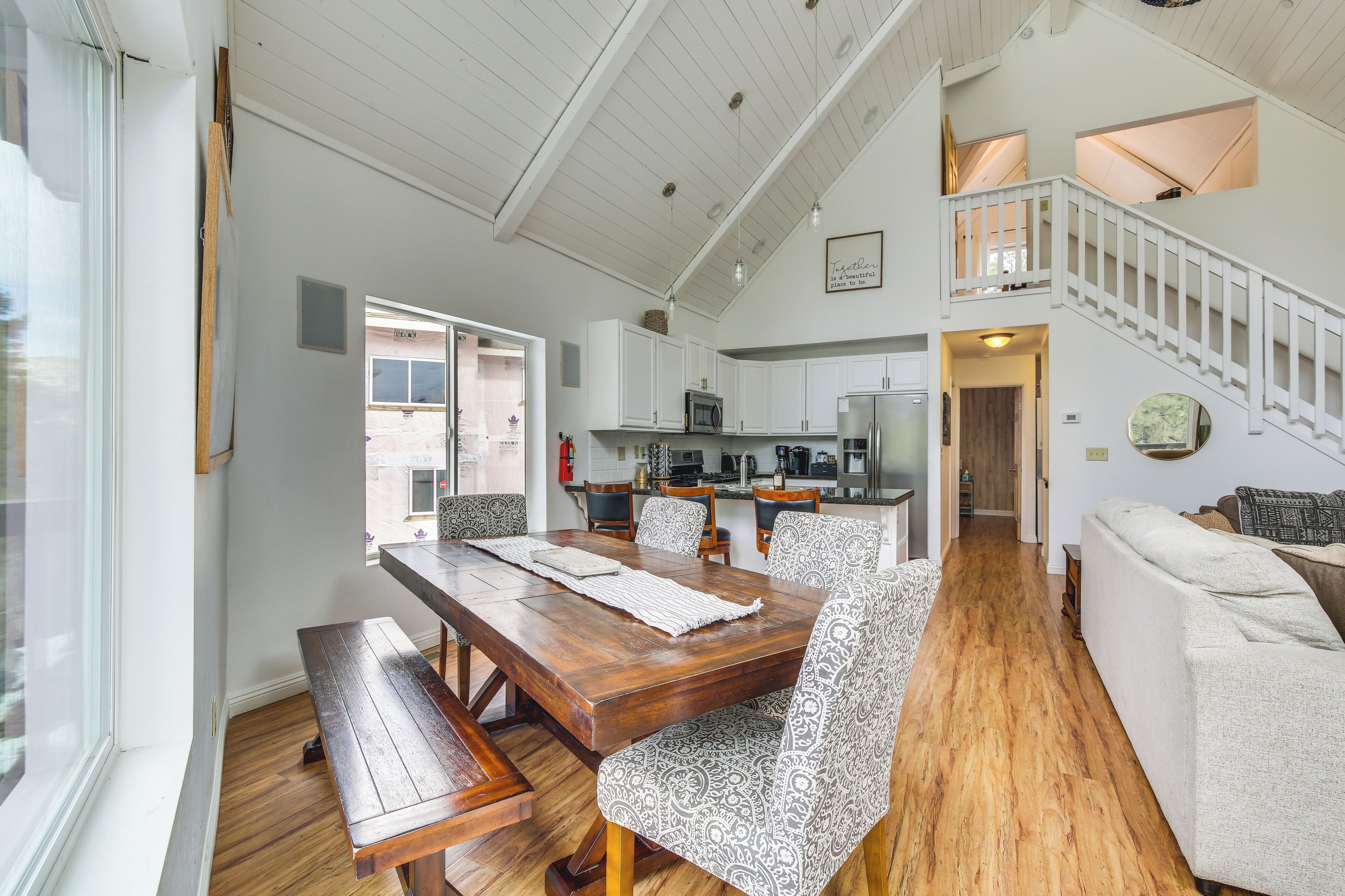 A spacious living area with a wooden dining table, patterned chairs, a white sofa, and a staircase leading to an upper level.