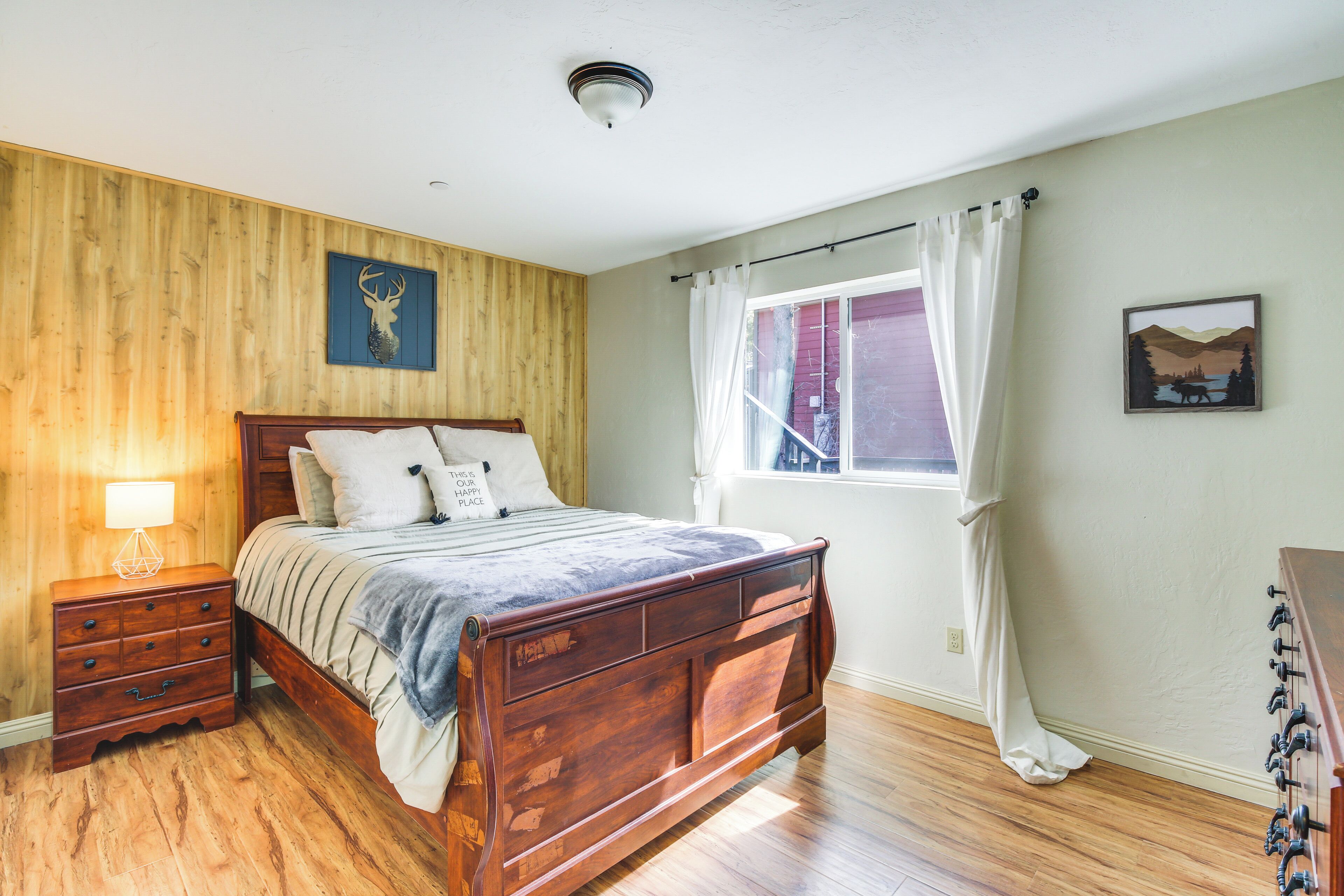 A bedroom with a wooden bed, a nightstand, a window with curtains, and a wall-mounted lamp.