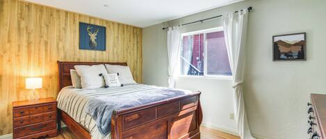 A bedroom with a wooden bed, a nightstand, a window with curtains, and a wall-mounted lamp.