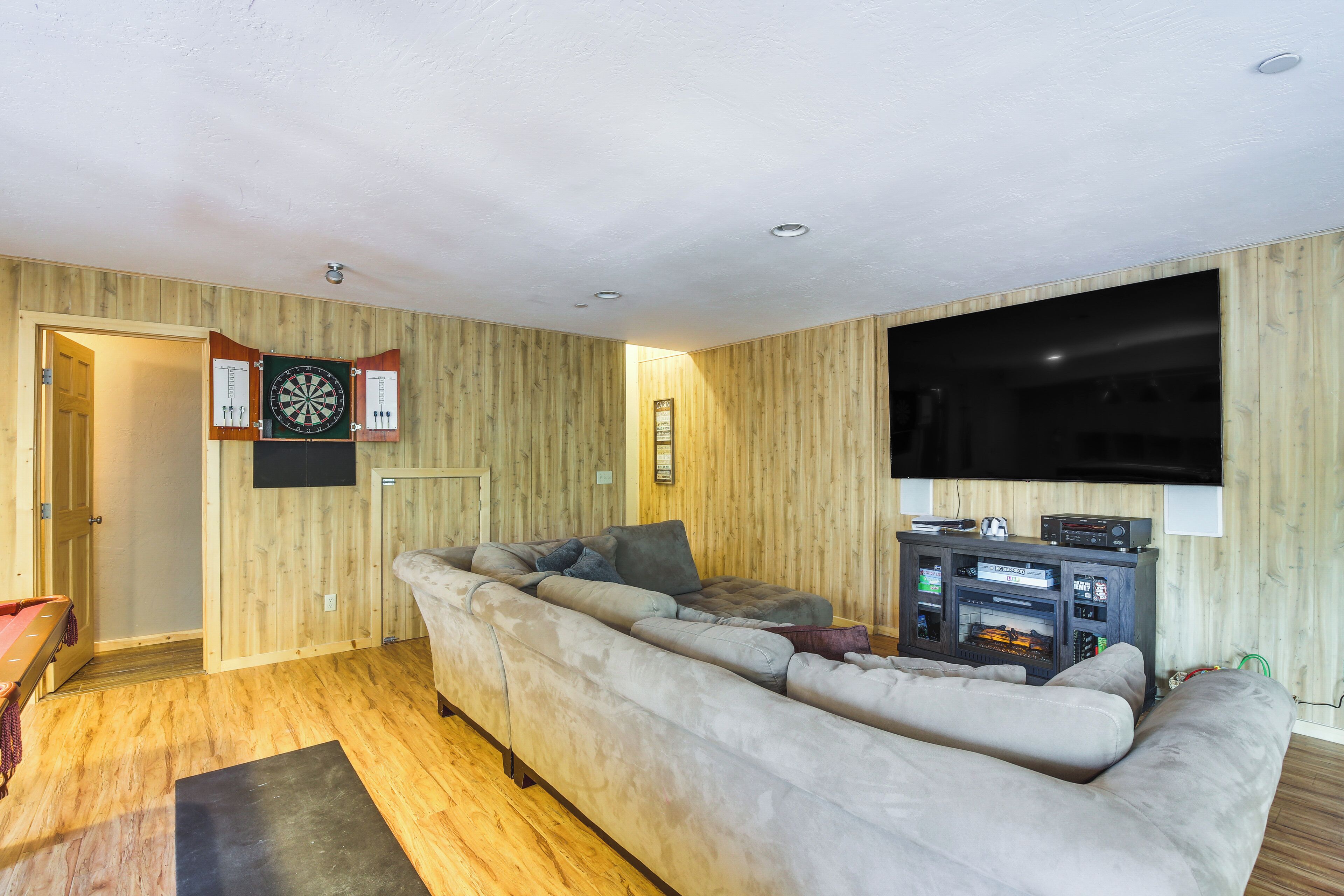 Un salón con un sofá seccional grande de color gris, un televisor de pantalla plana, paredes de madera y un dardo en la pared.