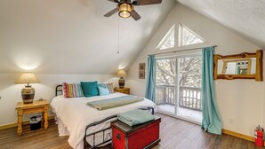 A bedroom with a sloped ceiling, a bed with multiple pillows, a nightstand with a lamp, a window with curtains, and a red trunk. - Mountain Home w/ Deck: 1 Mi to Angel Fire Resort! (Angel Fire)