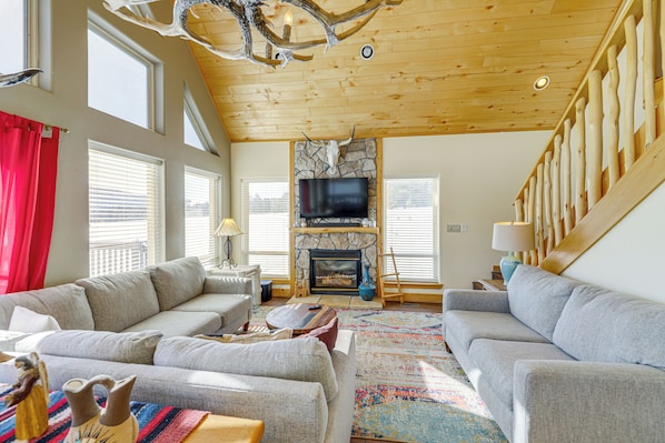 A spacious living room with a stone fireplace, a large flat-screen TV, and a wooden ceiling. - Mountain Home w/ Deck: 1 Mi to Angel Fire Resort! (Angel Fire)