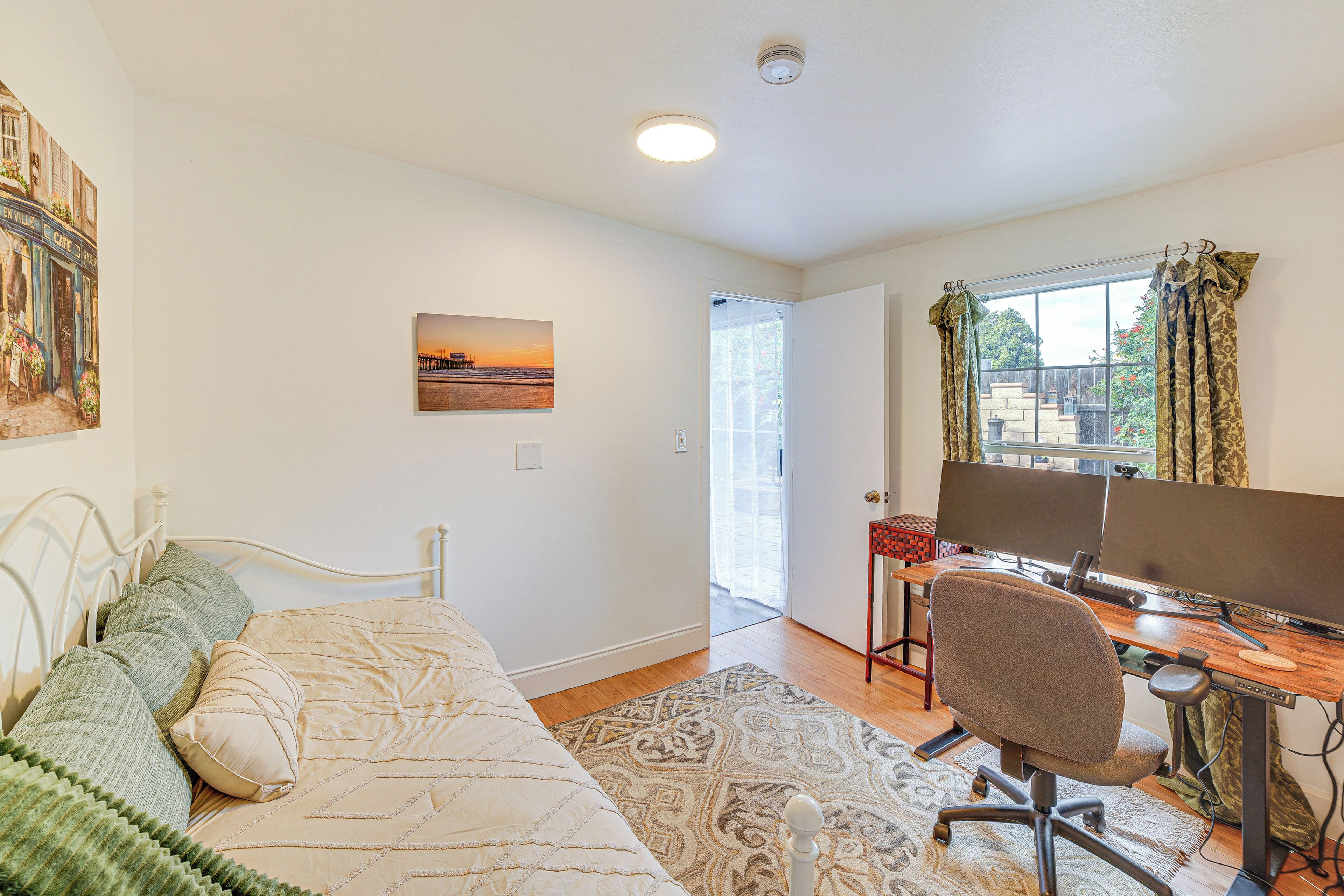 A bedroom with a bed, a desk with a computer, a window with curtains, and a door leading to another room.