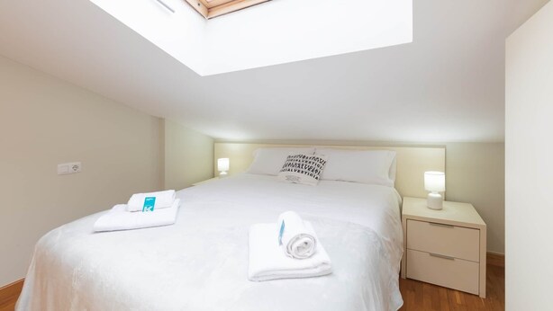 A hotel room with a skylight, a bed with white linens, a nightstand with a lamp, and a wall with a text message. - Ekain - baskeyrentals (Zestoa)
