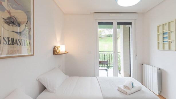 Une chambre à coucher avec un lit, une lampe de chevet, un tableau encadré et une vue sur l’extérieur.