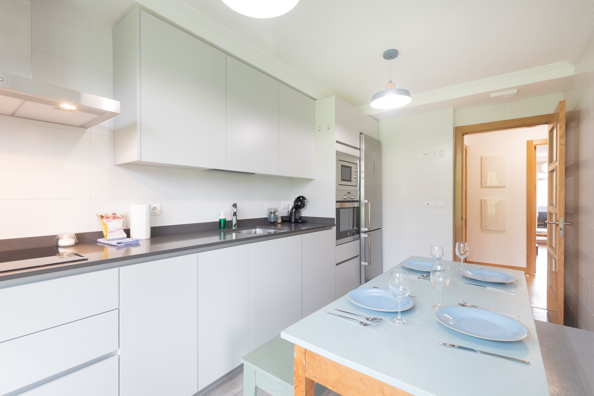 A modern kitchen with white cabinets, a stainless steel refrigerator, and a dining area set for four with blue plates and wine glasses.
