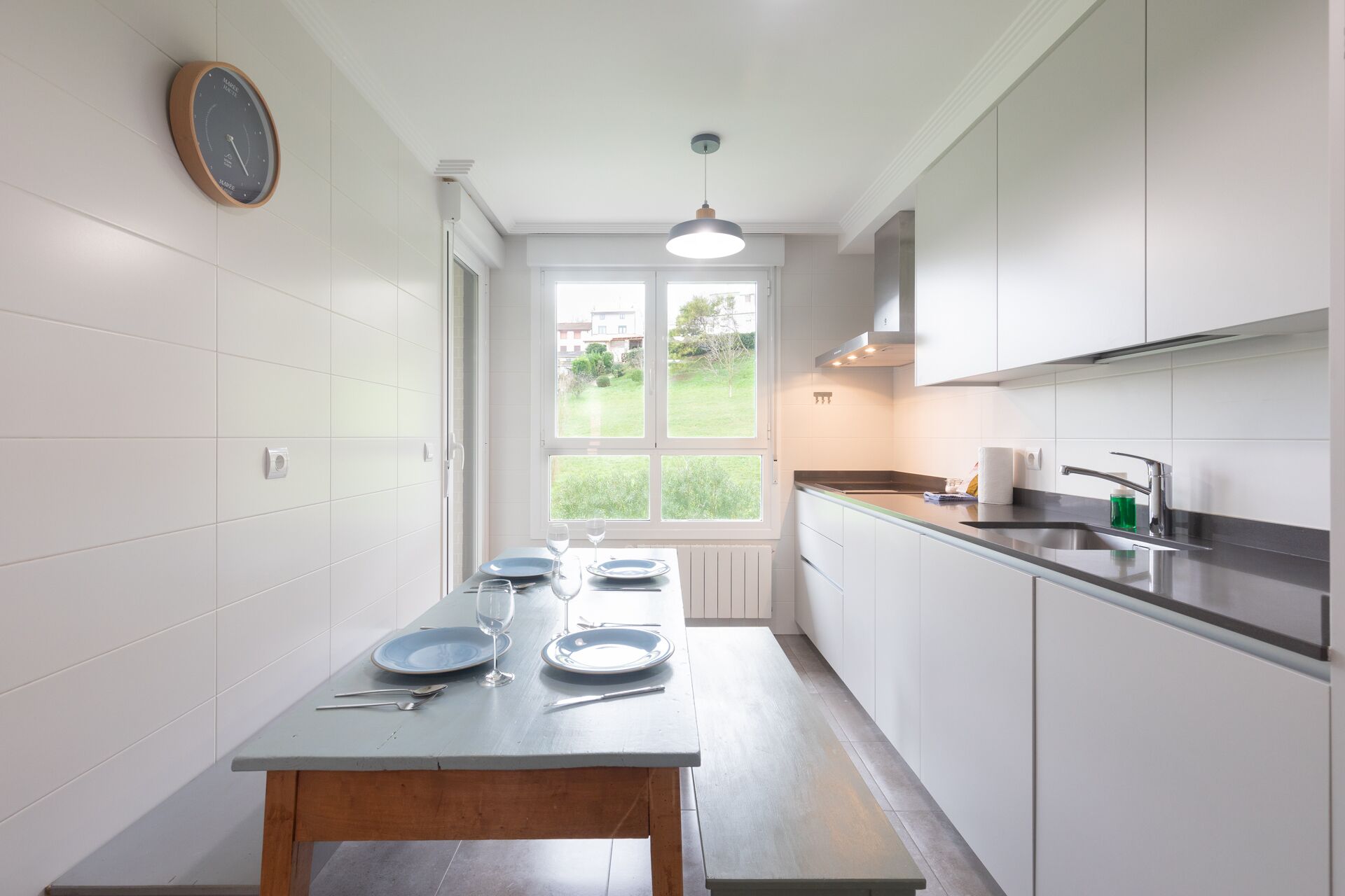 A modern kitchen with white cabinets, a wooden table set with plates and cutlery, a window with a view of greenery, and a clock on the wall.