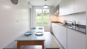 Une cuisine moderne avec des meubles blancs, une table en bois mise avec des assiettes et des couverts, une fenêtre donnant sur un paysage verdoyant et une horloge au mur.