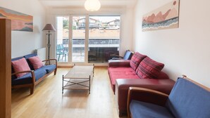 Un salon avec un canapé, un fauteuil et une petite table. On peut voir l’extérieur à travers une porte vitrée.