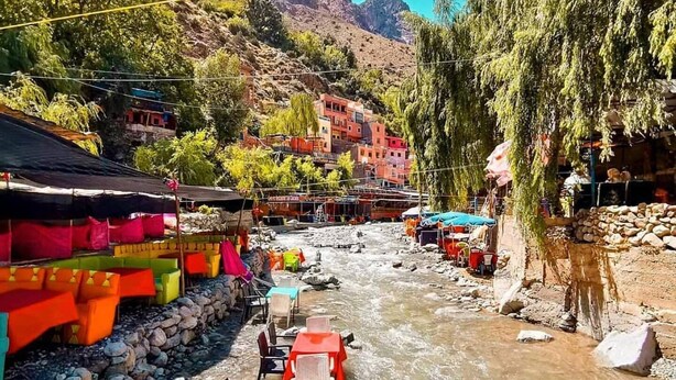 Un restaurante a la orilla del río con terraza, vistas a montañas y edificios coloridos al fondo.