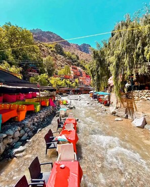 Un restaurante a la orilla del río con terraza, vistas a montañas y edificios coloridos al fondo.