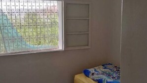 Una habitación pequeña con una cama individual, una ventana con barrotes y una pared blanca con un estante.