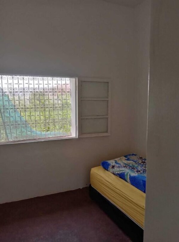 Una habitación pequeña con una cama individual, una ventana con barrotes y una pared blanca con un estante.