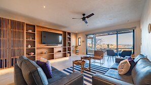 A modern living room with a large flat-screen TV, a ceiling fan, and a large window offering a view of the outdoors. - Private Oceanfront Rooftop Plunge Pool & Beach Access (Huatulco)