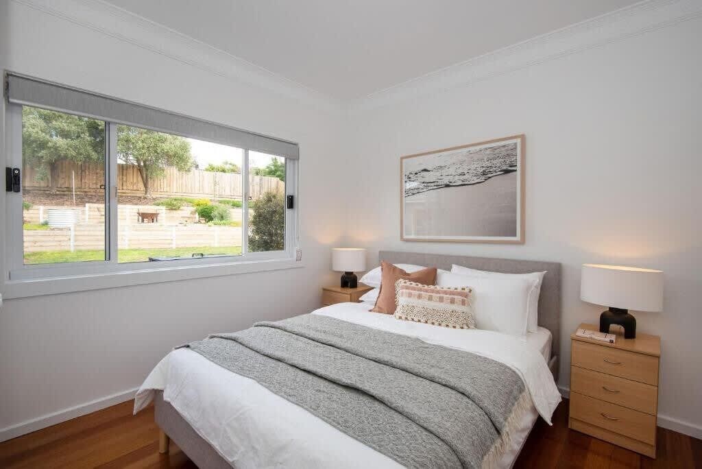 A bedroom with a large window, a bed with white bedding, two bedside tables with lamps, and a framed picture on the wall.