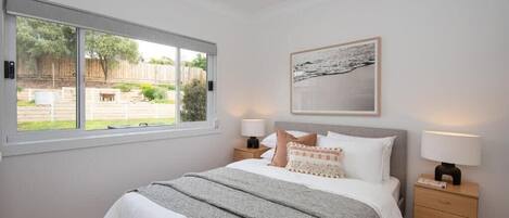 A bedroom with a large window, a bed with white bedding, two bedside tables with lamps, and a framed picture on the wall.