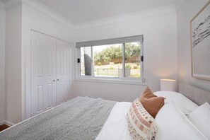 A bedroom with a large window, white walls, and a bed with white and brown bedding.