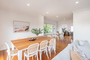 A spacious dining area with a wooden table and white wicker chairs, a large window, and a view of greenery outside.