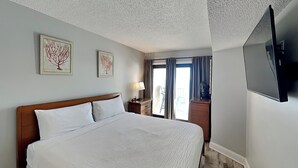 A hotel room with a large bed, a flat-screen TV, two framed artworks, a nightstand with a lamp, and a view of the outdoors through a sliding door.