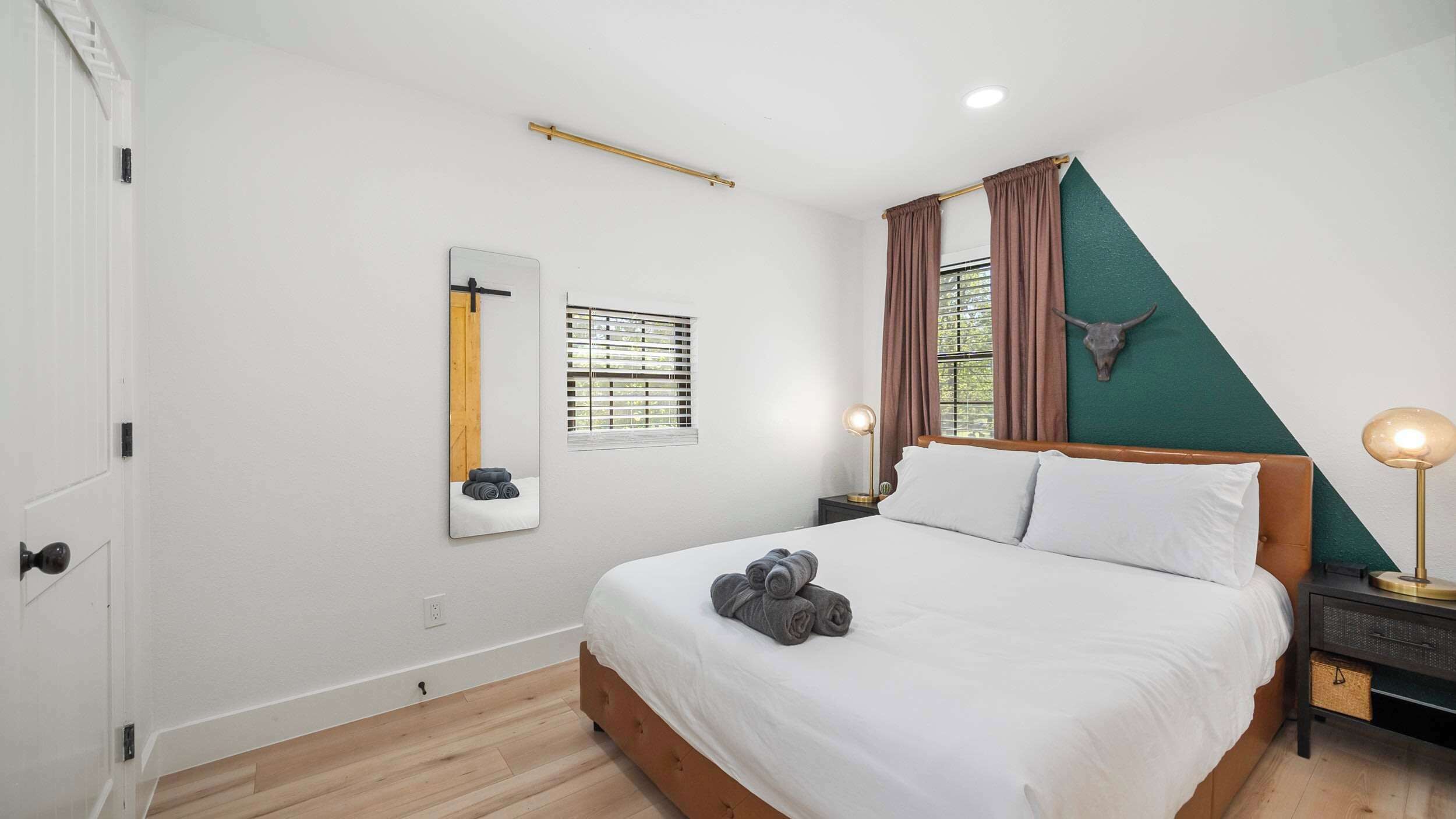 A bedroom with a bed, a bedside lamp, a mirror, a window with curtains, and a wall-mounted bull skull decoration.