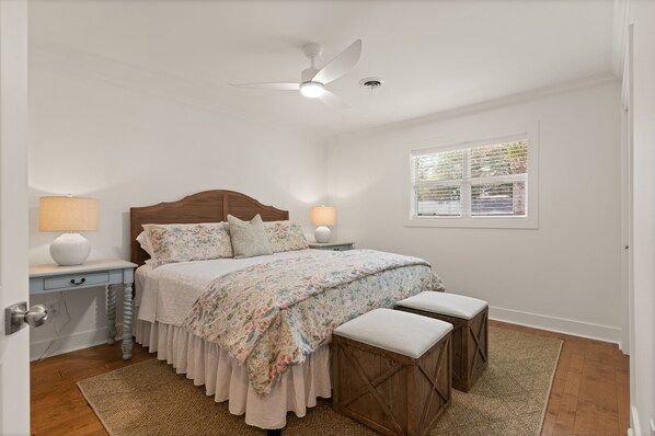 Une chambre à coucher comprenant un lit, deux tables de chevet avec des lampes, un ventilateur de plafond, une fenêtre avec des stores et un banc en bois.