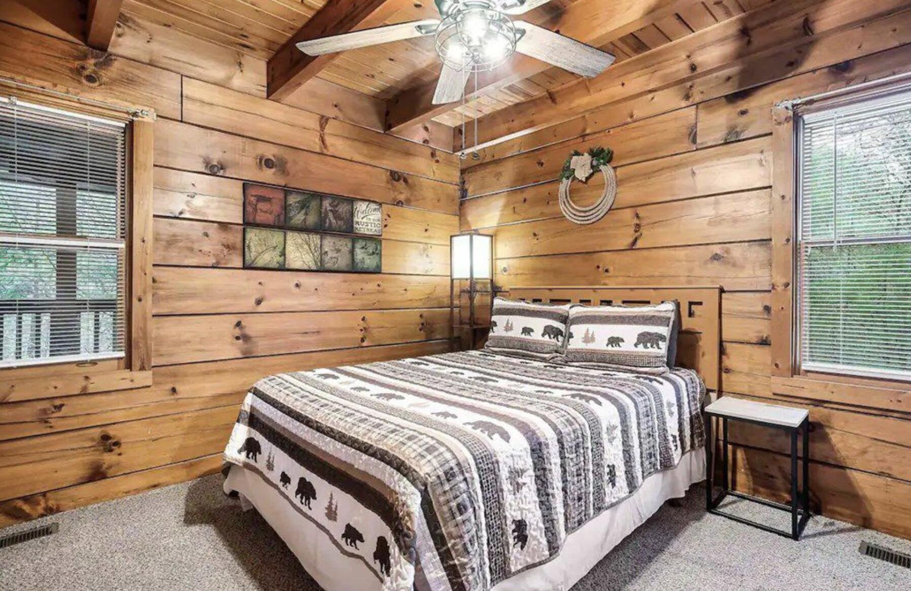 A wooden cabin bedroom with a bed, a ceiling fan, and a window with blinds.